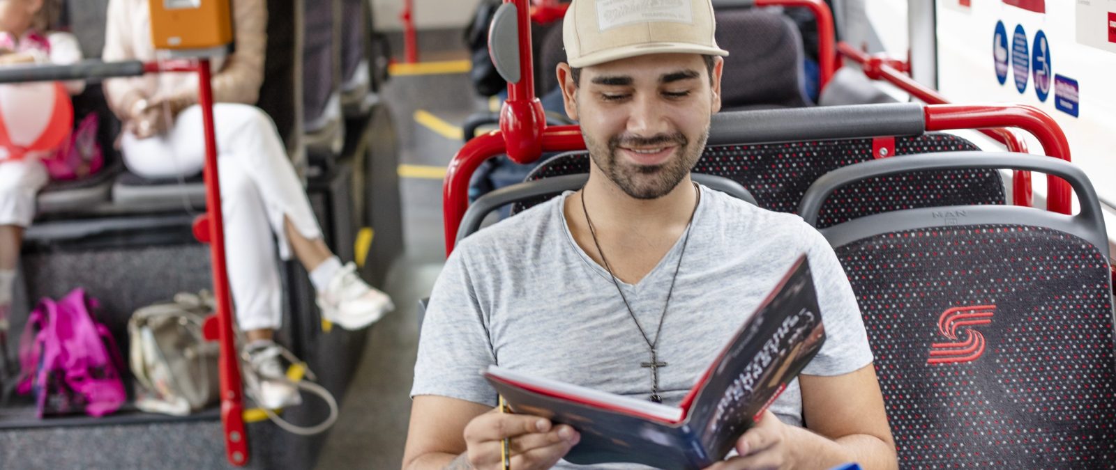 Junger Mann mit Baseball-Cap sitzt im Bus und hält ein Buch in der Hand