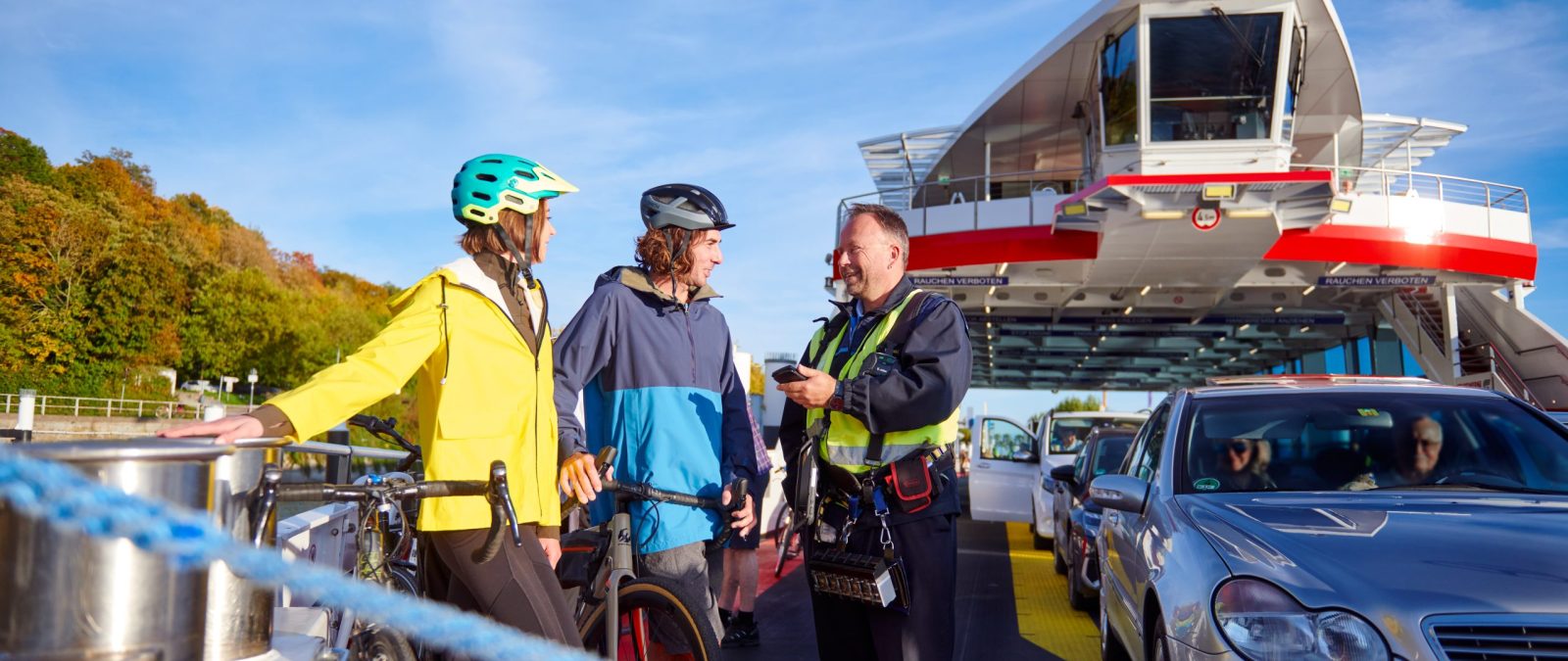 Zwei Personen mit Fahrrädern unterhalten sich mit einem Kassiermatrosen auf einem Fährschiff.